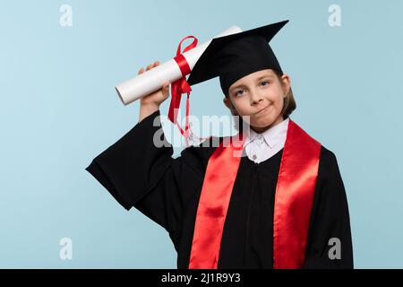 Ragazza di 9-11 anni con cappuccio di laurea e accappatoio per cerimonie con certificato di diploma su sfondo azzurro. Laureato per celebrare la laurea. Concetto di istruzione. Scuola elementare di successo Foto Stock