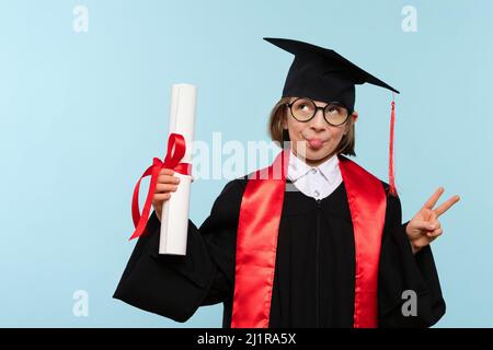 Ragazza di 9-11 anni con cappuccio di laurea, occhiali rotondi e accappatoio da cerimonia con certificato di diploma su sfondo blu. Il bambino fa un grimaces, mostra la lingua ed il segno di pace Foto Stock