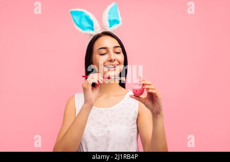 Donna felice in orecchie di coniglio dipinge uova di pasqua, preparazione per la festa della pasqua, in studio su sfondo rosa, cristianesimo Foto Stock