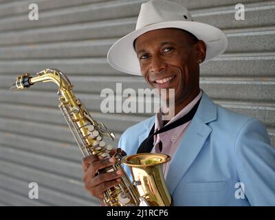 Bel giovane ed elegante sassofonista afro-cubano con cappello Panama bianco tiene il suo sassofono lucido lucido e sorride per lo spettatore. Foto Stock