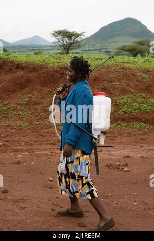 Una donna cammina trasportando un'irroratrice a mano sulla strada principale vicino al sito del progetto IPI che è diventata impraticabile per le auto dopo la pioggia pesante. Foto Stock