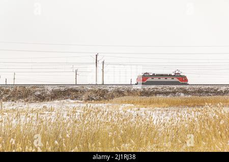 Un trenino elettrico solato senza vagoni si muove lungo la ferrovia passando l'erba gialla sullo sfondo della neve bianca. Foto Stock