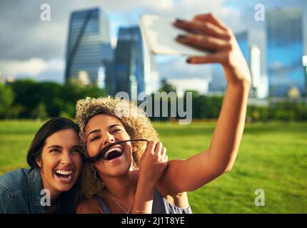 Ci piace immortalare i nostri stupidi momenti. Scatto corto di due giovani amiche in posa per un selfie insieme in un parco. Foto Stock