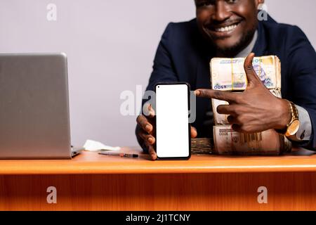 Un giovane nero gioioso seduto in ufficio con i pacchetti di denaro, un computer portatile che mostra il suo schermo del telefono Foto Stock