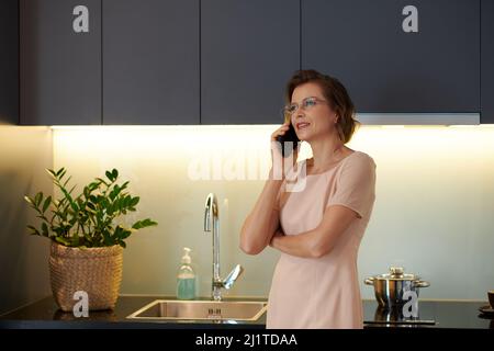 Ritratto di donna di mezza età positiva in piedi al banco della cucina e chiamare un amico per discutere le notizie Foto Stock