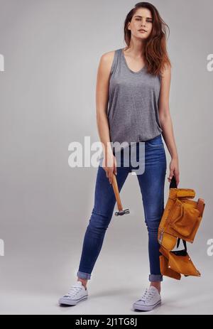 Posso risolvere qualsiasi problema. Ritratto completo in studio di una giovane donna vestita casualmente che tiene un martello e una cintura portautensili. Foto Stock