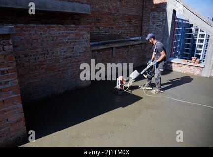 Vista laterale dell'operatore in condizioni di lavoro uniformi con l'attrezzatura e livellamento del pavimento in condizioni meteorologiche favorevoli. Concetto di costruzione di processo nuova casa moderna e fare muro. Foto Stock