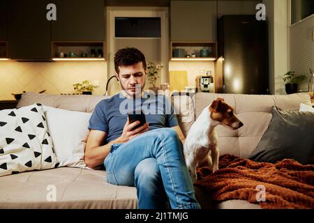Uomo che riposa sul divano con il suo cane e usando lo smartphone Foto Stock