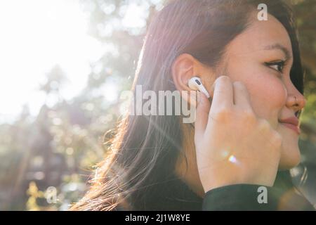Vista laterale di una femmina asiatica sorridente con le moderne cuffie wireless per ascoltare la musica nel parco soleggiato Foto Stock