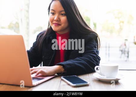 Coltivare giovane donna asiatica telelavoratore seduto al tavolo con una tazza di bevanda calda e smartphone e lavorando su netbook durante la giornata di lavoro online Foto Stock
