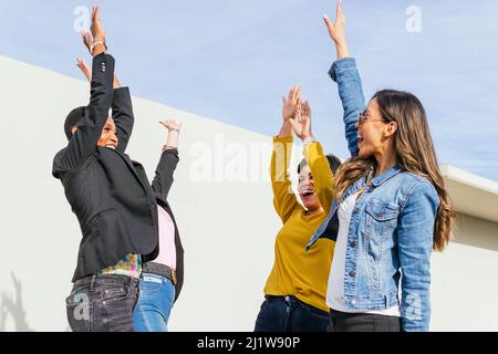 Gruppo di amici femminili eccitati che alzano le braccia e si guardano mentre si alza in piedi sulla strada soleggiata della città Foto Stock