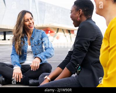 Gruppo completo del corpo di amici femminili multirazziali che si guardano a vicenda e chattando mentre si siede su scandole tetto in metallo vicino ostello in città Foto Stock