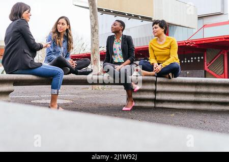 Gruppo completo del corpo di amici femminili multirazziali che si guardano a vicenda e chattando mentre si siede su scandole tetto in metallo vicino ostello in città Foto Stock