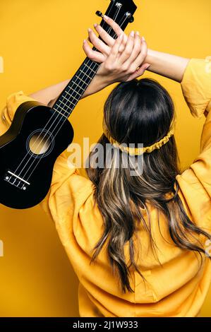 vista posteriore del musicista femminile irriconoscibile in abbigliamento luminoso che alza l'ukulele acustico per rompere in studio su sfondo giallo Foto Stock