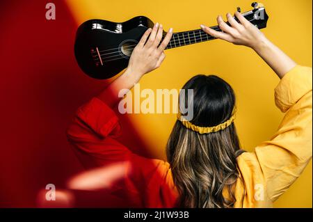vista posteriore del musicista femminile irriconoscibile in abbigliamento luminoso che alza l'ukulele acustico per rompere in studio su sfondo giallo Foto Stock