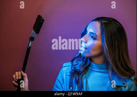 Vista laterale di giovane donna attraente indossando il ponticello e la giacca di cuoio che tiene la luce blu LED e il monopiede Foto Stock