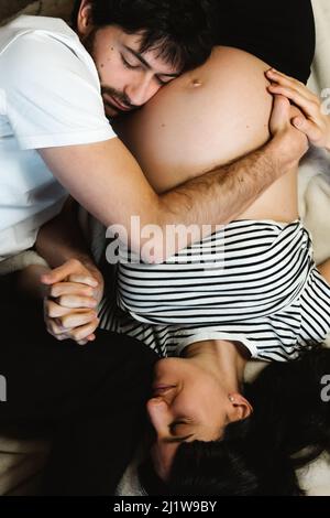 Vista dall'alto di un dolce marito che abbraccia il ventre della moglie incinta mentre si rilassa sul letto morbido di casa Foto Stock