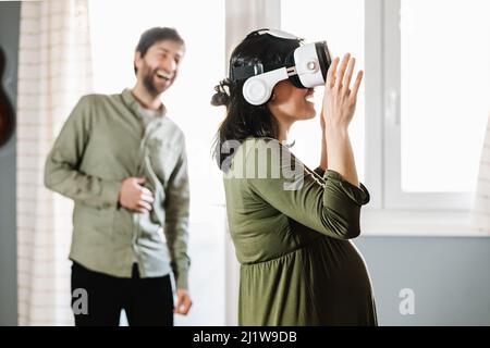 Stupito giovane donna incinta ispanica con capelli scuri in vestito guardando lontano mentre esplorare il cyberspazio in moderni occhiali VR in piedi a casa vicino smilin Foto Stock