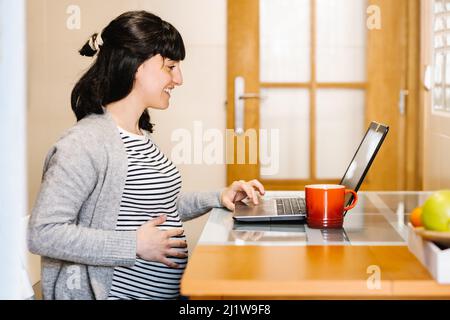 Vista laterale della giovane ragazza ispanica femminile incinta positiva freelancer con capelli scuri sorridenti e toccando il ventre mentre si lavora da remoto sul portatile al tavolo a. Foto Stock
