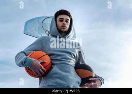 Da sotto di uno sportivo ispanico serio con basketball guardando la macchina fotografica mentre si trova vicino a basket hoop sul terreno sportivo durante l'allenamento su st Foto Stock