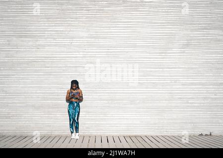 Vista frontale pieno corpo di felice giovane afroamericana femmina con dreadlock indossando abbigliamento sportivo ascoltare la musica in auricolari e navigare cellulare Foto Stock