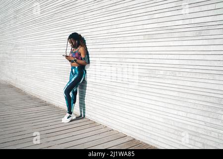 Giovane donna afroamericana con dreadlock indossando abbigliamento sportivo ascoltando la musica in auricolari e navigando cellulare mentre si appoggia sulla parete di legno d Foto Stock