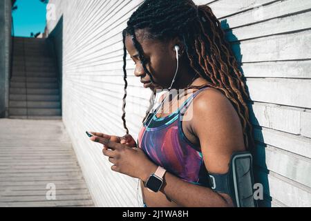 Giovane donna afroamericana con dreadlock indossando abbigliamento sportivo ascoltando la musica in auricolari e navigando cellulare mentre si appoggia sulla parete di legno d Foto Stock