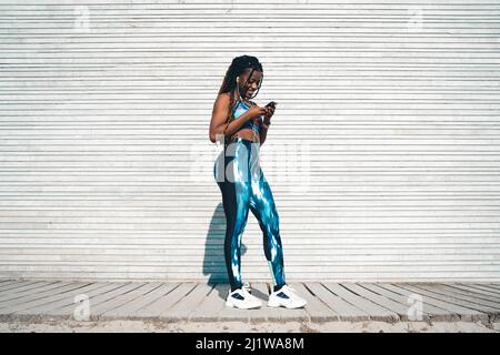 Vista laterale della giovane donna afroamericana con dreadlock indossando abbigliamento sportivo ascoltando la musica nelle cuffie e navigando sul cellulare mentre si appoggiano Foto Stock