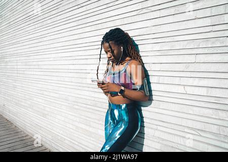 Giovane donna afroamericana con dreadlock indossando abbigliamento sportivo ascoltando la musica in auricolari e navigando cellulare mentre si appoggia sulla parete di legno d Foto Stock