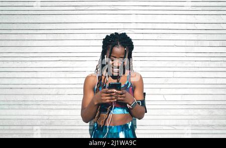Vista frontale della giovane ragazza afro-americana felice con dreadlock indossando abbigliamento sportivo ascoltare musica in auricolari e navigare cellulare mentre magro Foto Stock