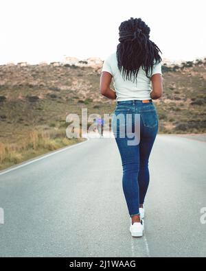Vista posteriore di anonima e pacifica femmina afroamericana che cammina lungo la strada asfaltata negli altopiani Foto Stock