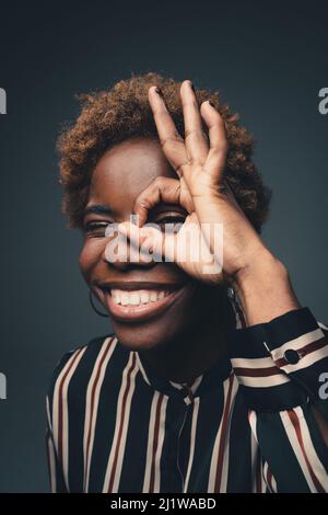 Felice bambina afroamericana con capelli ricci corti sorridendo ampiamente e mostrando il simbolo giusto intorno all'occhio in studio scuro Foto Stock