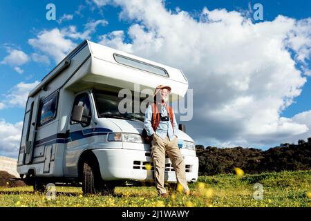 Corpo pieno di turista maschile anziano in cappello in piedi vicino auto RV parcheggiata su campo erboso mentre osservano la natura il giorno d'estate Foto Stock