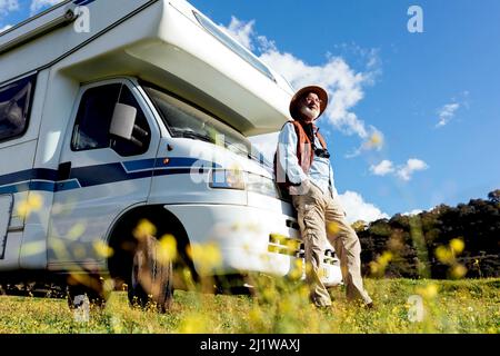 Corpo pieno di turista maschile anziano in cappello in piedi vicino auto RV parcheggiata su campo erboso mentre osservano la natura il giorno d'estate Foto Stock