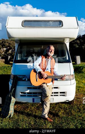 Il viaggiatore anziano di talento bearded maschio che suona la canzone sulla chitarra acustica mentre si alza vicino ad un'automobile di RV durante il viaggio di strada nella natura Foto Stock