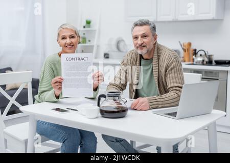 I pensionati che hanno contratto e guardano la macchina fotografica vicino a dispositivi e caffè in cucina Foto Stock