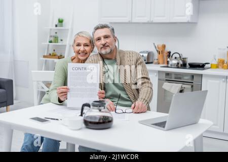 Sorridente donna anziana che detiene un contratto vicino a marito e dispositivi in cucina Foto Stock