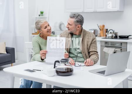 Donna anziana allegra che tiene contratto e che guarda il marito vicino a gadget e caffè in cucina Foto Stock