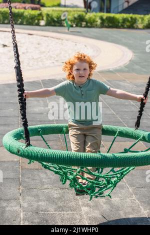 ragazzo curly che guarda la macchina fotografica mentre si diverte in amaca sul parco giochi Foto Stock