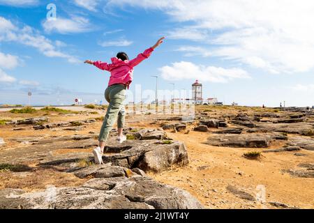 Ragazza che salta su rocce con le braccia allungate a capo Carvoeiro con il faro sullo sfondo Foto Stock