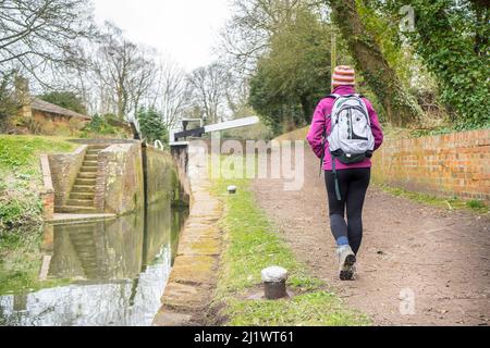 Vista posteriore della donna isolata in cappello stripy che porta zaino sul retro, camminando lungo il sentiero di un canale rurale del Regno Unito verso porte a serratura aperte e gradini. Foto Stock