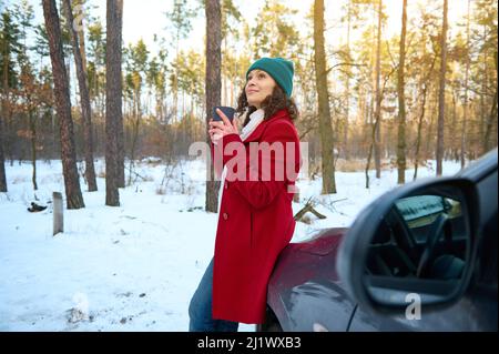 Donna viaggiatore tiene thermo tazza con tè caldo, gode di pausa caffè all'aperto, seduto sul cofano auto in nevoso bosco. Raggi del sole che cadono sulla foresta p Foto Stock