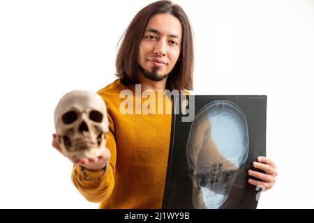 Uomo caucasico con capelli lunghi che tiene in mano il cranio artificiale e il cranio radiografia mentre guarda la fotocamera. Studio girato su sfondo bianco. Foto di alta qualità Foto Stock