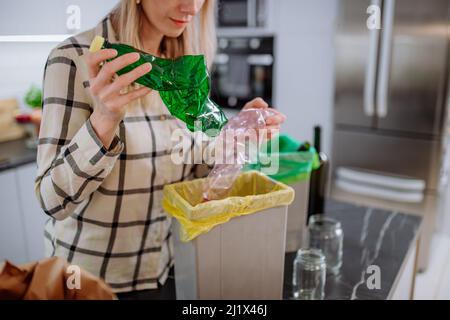 Sezione centrale della donna gettando una bottiglia di plastica vuota in un contenitore di riciclaggio in cucina. Foto Stock