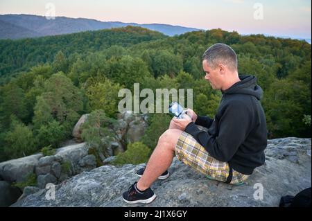 Uomo che utilizza il drone tramite telecomando. Uomo che utilizza il drone al tramonto per foto e video making mentre si siede in cima al masso alto in montagna. Foto Stock