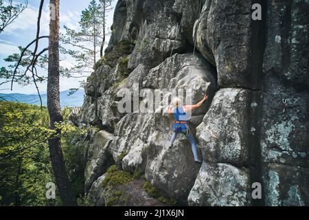 Donna di alto grado che si arrampica su un percorso difficile su un alto masso con corda. Arrampicata su roccia senza paura nella giornata estiva. Concetto di sport estremo, avventure e stile di vita attivo. Vista aerea. Foto Stock