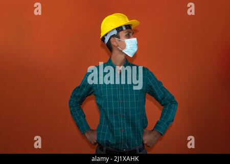 Ingegnere, architetto o lavoratore con casco giallo e maschera pandemica isolato su sfondo arancione. Lavoratore con casco giallo e maschera pandemica isolato Foto Stock
