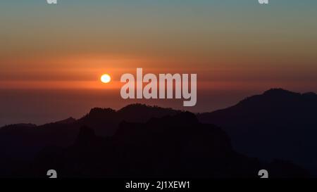 Tramonto mozzafiato sulle aspre montagne dell'isola di Gran Canaria, Isole Canarie, Spagna con sole rosso nel cielo colorato e le silhouette delle colline. Foto Stock