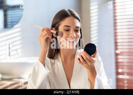 Risveglio giornaliero. Giovane donna sorridente che applica il Blush con la spazzola di trucco in bagno Foto Stock