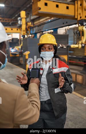 Donna africana in hardhat e uniforme indossare maschera protettiva parlare con giornalista durante il reportage in fabbrica Foto Stock
